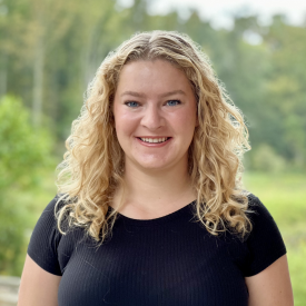 A woman with curly blonde hair and a black t-shirt smiling, in front of a forest.