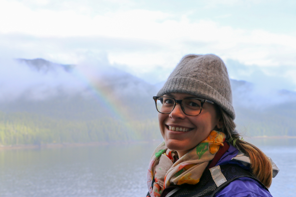 A woman--Dr. Linsey Haram--in a light gray beanie, glasses, and a scarf. This photo was taken above the shoulders, and Dr. Haram is smiling, facing the camera. The background is more faded and out of focus, but shows mountains in the distance, behind clouds, and part of a rainbow reaching the ground. Below the mountains is a forest and in front of that is a body of water