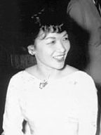 A middle-aged lady wearing a light-colored dress and with short hair, fluffy at the front, sits at a dinner table smiling.