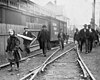 A girl in old-fashioned winter clothing, in foreground, and several adult men wearing open winter coats and hats, in background. All are walking along railroad tracks and toward the viewer. Beyond the people are a wooden fence, a railroad box car, and a large building.