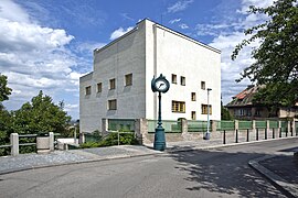 Vue d'une maison particulière de taille importante, cubique et ajouré de quelques fenêtres jaunes. La rue et une horloge publique sont au premier plan.