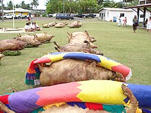 Photo en couleur montrant des cochons cuits, disposés régulièrement sur le sol les pattes en l'air. Des nattes colorées sont posées sur certains d'entre eux. Des voitures sont visibles à l'arrière plan, quelques personnes marchent sur le côté. Un groupe de personnes est assis sur une natte sur le côté gauche de la photo.