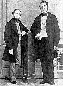 Black-and-white image of two middle-aged men, either one leaning with one elbow on a wooden column in the middle. Both wear long jackets, and the shorter man on the left has a beard.