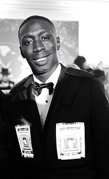 black-and-white image of Khaby Lame wearing a dark suit with bowtie, grinning at camera