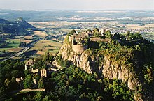 Ruins of a fortress on a volcanic mountain, overlooking a small city, farmlands, and, in the background, a lake.