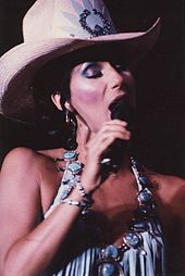 Cher sings into a microphone, wearing a wide-brimmed rhinestone-studded cowboy hat and a revealing outfit accented with large turquoise jewelry.