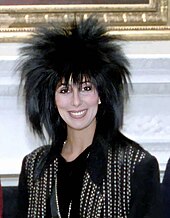 Cher smiles in a black outfit with metallic studs, sporting a voluminous, spiky hairstyle.