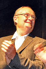 A portrait of a bespectacled, balding male in his late fifites. He is wearing a brown coat over a white collared shirt.