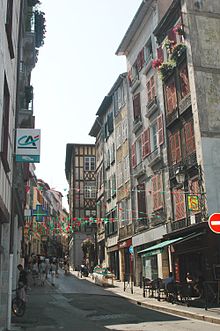 Vue d’une rue de bâtiments de 3 et 4 étages, aux façades blanches à pans de bois peints.