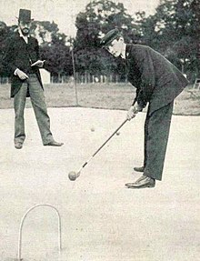 Photo d'un homme poussant la boule en direction d'un arceau. À l'arrière se trouve un homme en costume tenant un carnet.