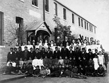 Posed, group photo of students and teachers, dressed in black and white, outside a brick building in Regina, Saskatchewan