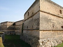 Uma estrutura de pedra de três andares com paredes lisas e uma base ásperamente cortada. As paredes são angulares e têm aberturas.