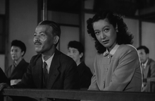 A theatre at which a Noh play is being performed: Prof. Somiya is wearing a business suit and tie and Noriko is wearing a simple, Western-style dress with a collar; Shukichi is looking straight ahead towards the left frame of the picture, smiling, and Noriko, not smiling, is looking toward the right frame of the picture towards an unseen person.