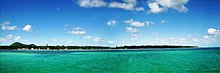 Panorama shot of an island covered in trees, in front of a clear greenish blue sea and sunny weather