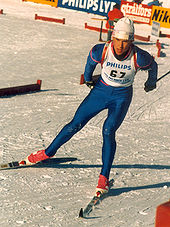 A man skies on a snow-covered course. He is wearing a white vest on top of a blue skin-tight jumpsuit, a white winter cap, and red ski boots. On his back, he carries a rifle.