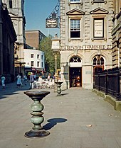 Two ornate metal pillars with large dishes on top in a paved street, with an eighteenth-century stone building behind, upon which can be seen the words "Tea Blenders Estabklishec 177-". People sitting at café-style tables outside. On the right are iron railings.