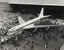 Photo noir et blanc. Un grand avion sort d'usine, entouré de nombreuses personnes.