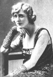 A white woman with coiffed hair, seated, wearing a dress with distinctive striped sleeves