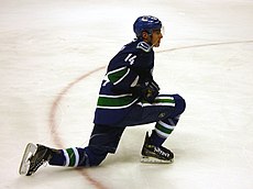 An ice hockey player dressed in a blue jersey with blue and green trim. He is stretching with one knee on the ice and the other lunged forward. He rests his stick on one leg while looking forward.