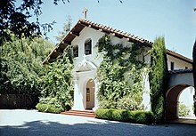 Color photograph of the front of a church showing tall plants flanking an arched entrance, and a gabled roof covered by mission-style curved tiles.