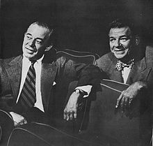 Two men in business suits sit in theatre seats amiably discussing what they are watching