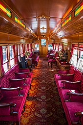 The ornate interior of the parlor consists of wooden walls and furniture, curtains, a rug covering the entire floor, and a vase filled with flowers on a table.