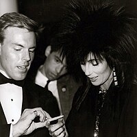 Cher, in a spiked hairstyle and dark outfit, signs an autograph while interacting with a man in a tuxedo.