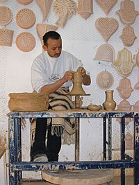 Potier de Guellala travaillant de l’argile sur son tour ; le mur derrière lui est couvert de poteries.