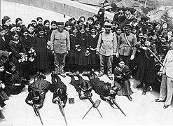 Photo de quatre écolières allongées sur le sol en train de s'entrainer au tir au fusil. Juste derrière elles, un groupe compact d'autres écolières observe la scène.