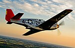 A single nosed-propeller plane is in mid flight above land. The red nose faces right and the underside of the plane is slightly in view. The plane has black propellers, grey base paint, and black letters reading A42. The 4 and 2 are separated by a United States roundel in black with a central white star. The roundel is also visible on the tops of the planes wings. The full length of the right side of the plane is visible.