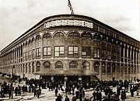 A black-and-white photograph of a large, imposing structure, viewed from outside, just above street level. Two vast walls, lined with windows, come from the viewer's left and right at 20-degree angles and meet at a rounded point, at which there is a covered entrance. The streets are crowded with people, many queuing to enter. Above the entrance neon lights spell the words "EBBETS FIELD" and above them an American flag flies on a pole.