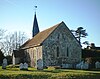 A low, broad, stone-built church with a tiled roof and a spire at the far end, with a porch projecting to the left