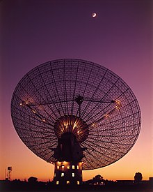 Photographie d'une antenne parabolique pointant vers le ciel, durant le crépuscule, la lune (croissant) étant visible sur le haut de l'image.