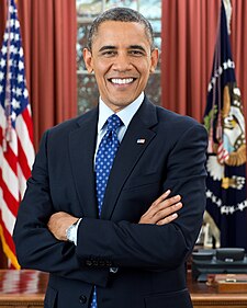 Barack Obama standing in front of a wooden writing desk and two flagpoles.