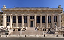 Frontale Farbfotografie eines klassizistischen Gebäudes mit einer breiten Säulenfassade und sechs antiken Steinfiguren in den Zwischenräumen. Am Treppenaufgang mit einem weißen Steingeländer sitzen zwei Löwenskulpturen und an beiden Seiten des Vorplatzes führen zwei weiße Steingeländer zu den Seitentüren. Das Gesims ist verziert und an beiden Dachenden stehen zwei Adlerfiguren.