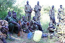 Photographie en couleurs montrant une dizaine d'hommes en treillis de camouflage, assis ou debout dans l'herbe entre des buissons, armes à la main. Deux bidons sont posés au centre de l'image.