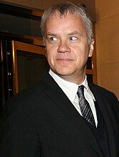 A standing caucasian man with short white and grey hair, wears glasses and a blue coat: He faces left towards the camera smiling.