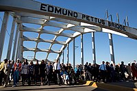 Large group of people walking together on bridge roadway under bridge arch saying "Edmund Pettus Bridge"