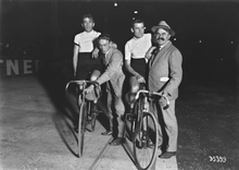 Photographie en noir et blanc montrant deux cyclistes montés sur leurs vélos, à l'arrêt sur une piste, s'appuyant sur deux hommes en civil.