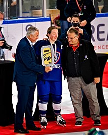 Joueuse de hockey debout avec son équipement sauf son casque, ses gants et sa crosse (ou bâton) qui reçoit un trophée de la main d'un homme à sa gauche. Un autre homme se tient à sa droite.