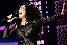 Cher sings on stage with one arm extended, wearing a black sheer bodysuit adorned with sparkling embellishments and dark curly hair cascading down.