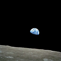 A black sky with a grey, cratered lunar horizon. A small blue Earth with scattered white clouds is just above the horizon, with about two-thirds of the Earth lit by the Sun and the remainder in darkness.