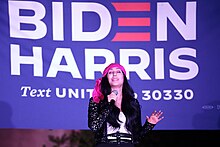 Cher sings on stage, wearing a pink headscarf and black sequined jacket, with a large "Biden Harris" banner in the background.