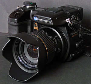 Oblique view of Konica Minolta DiMAGE A2 digital camera, which is a black camera with a prominent handgrip and lens, fitted with a petal-shaped lens hood. Lettering and an accent ring are colored gold.