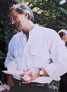 Jimmy is a tall man with short gray hair. He's holding a small party plate and smiling toward someone off-camera.