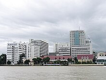 A large complex of buildings, most over ten storeys high, on the bank of a river; one bears a sign with the words "SIRIRAJ HOSPITAL"; another says "FACULTY OF NURSING"