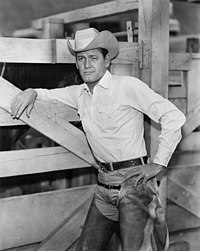 A man wearing a cowboy attire, leaning on a stable stall door.