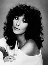 Cher poses with voluminous curly hair, wearing an off-the-shoulder white garment and gazing at the camera.