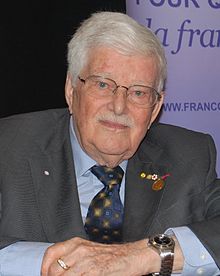 Photographie de Paul Gérin-Lajoie accoudé sur une table devant une bannière du Forum mondial de la langue française.