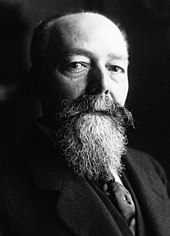 Portrait photographique en noir et blanc et de trois-quarts d'un homme à la chevelure et à la barbe grisonnantes, en veston-cravate